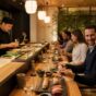 People dressed in smart casual clothes eating sushi at a modern restaurant with a sushi chef preparing food behind the counter.